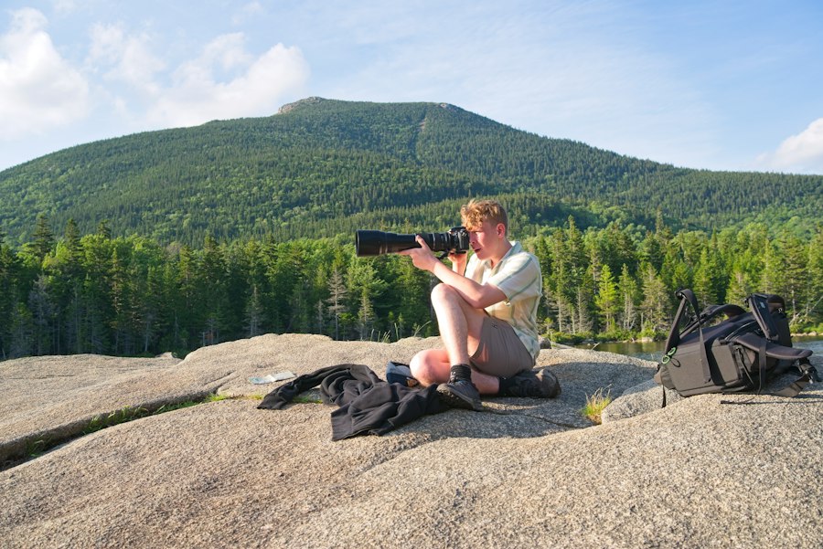 A person photographing a mountain landscape while sitting on a large granite rock